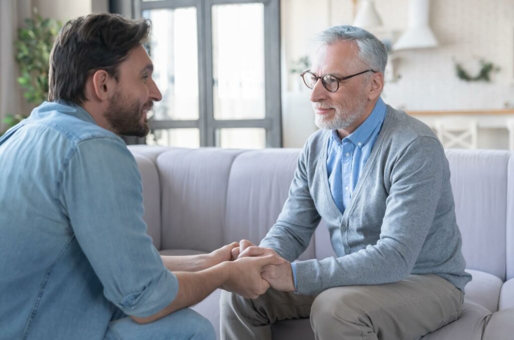 A smiling adult child sitting across from their older parent while holding their hands and asking them a question.