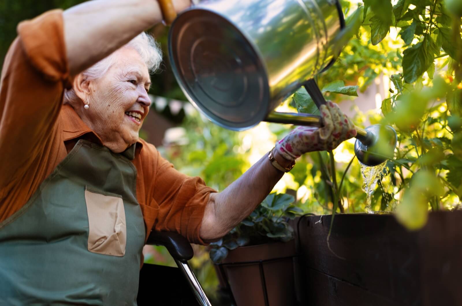 An older adult in assisted living sitting on a chair outdoors with a watering can, working independently in their garden.