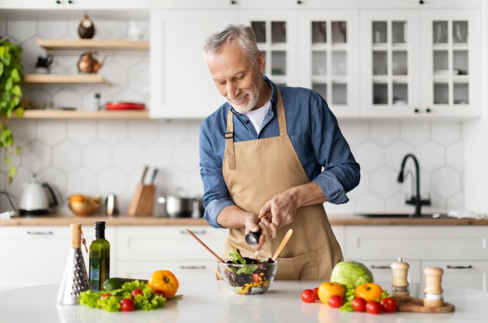 An older adult smiling while standing at an island in a brightly lit kitchen making a salad with various vegetables, olive oil, and seasonings, perfectly complementing their Mediterranean diet.