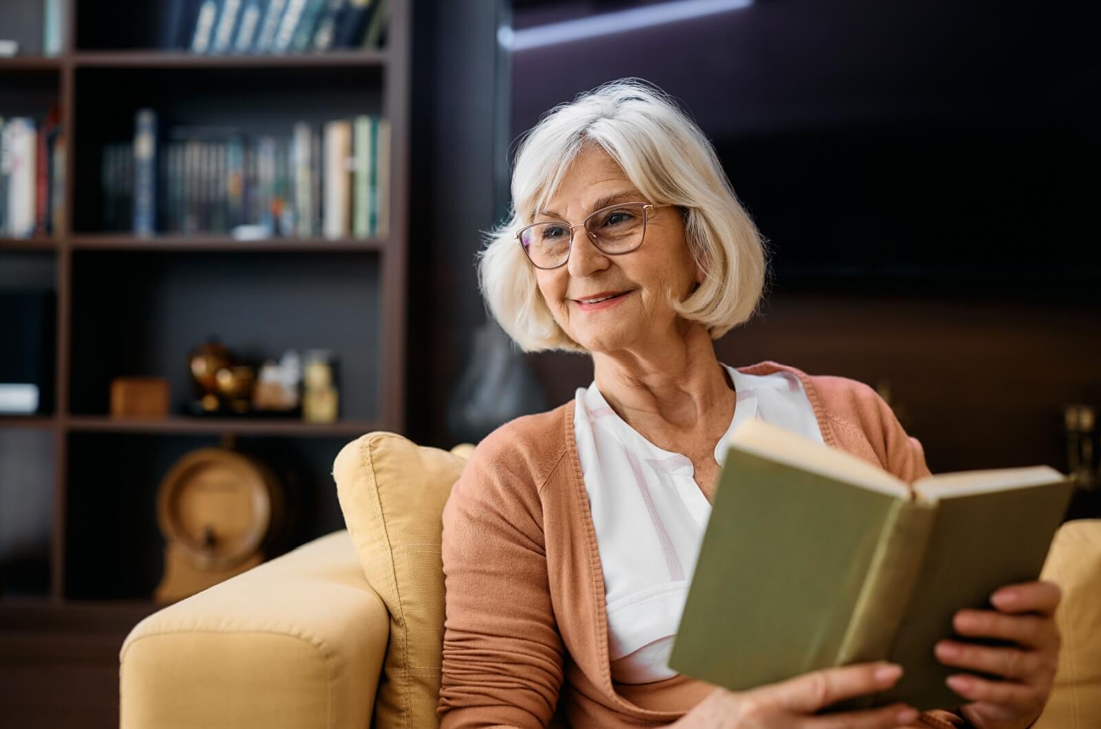 An older woman smiling and sitting on a couch holding a book open