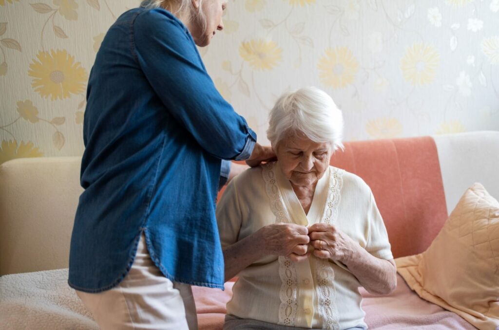 A caregiver helping an older adult sitting on a bed get dressed.