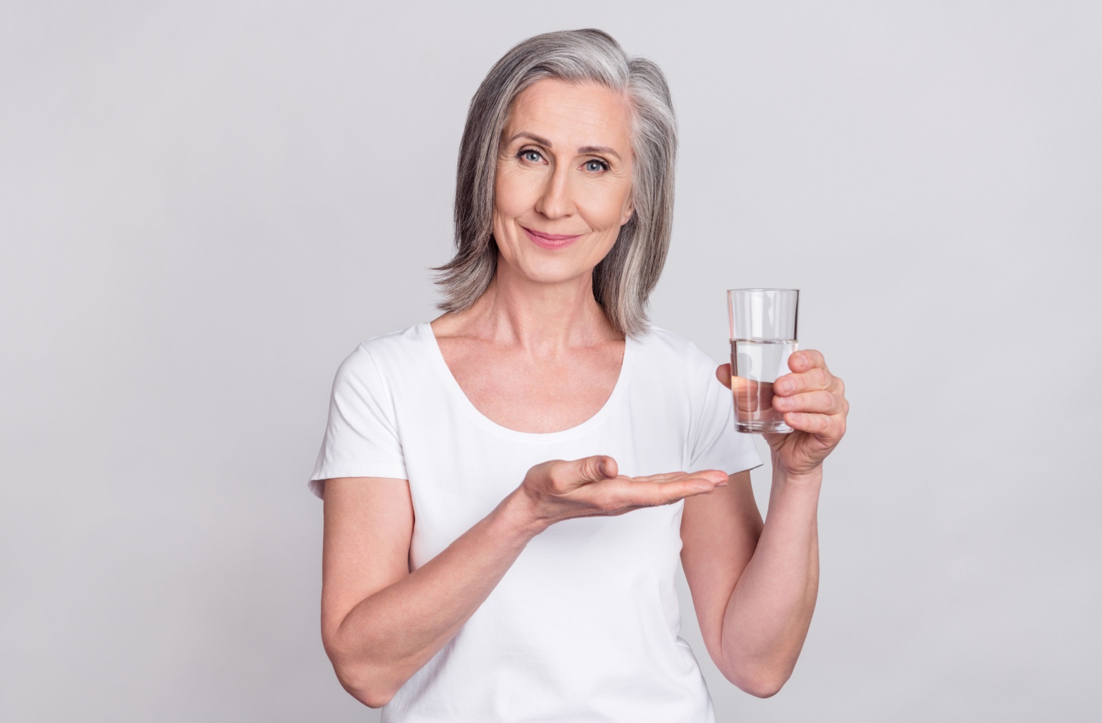 An older adult smiling and gesturing to a glass of water in front of a gray background.