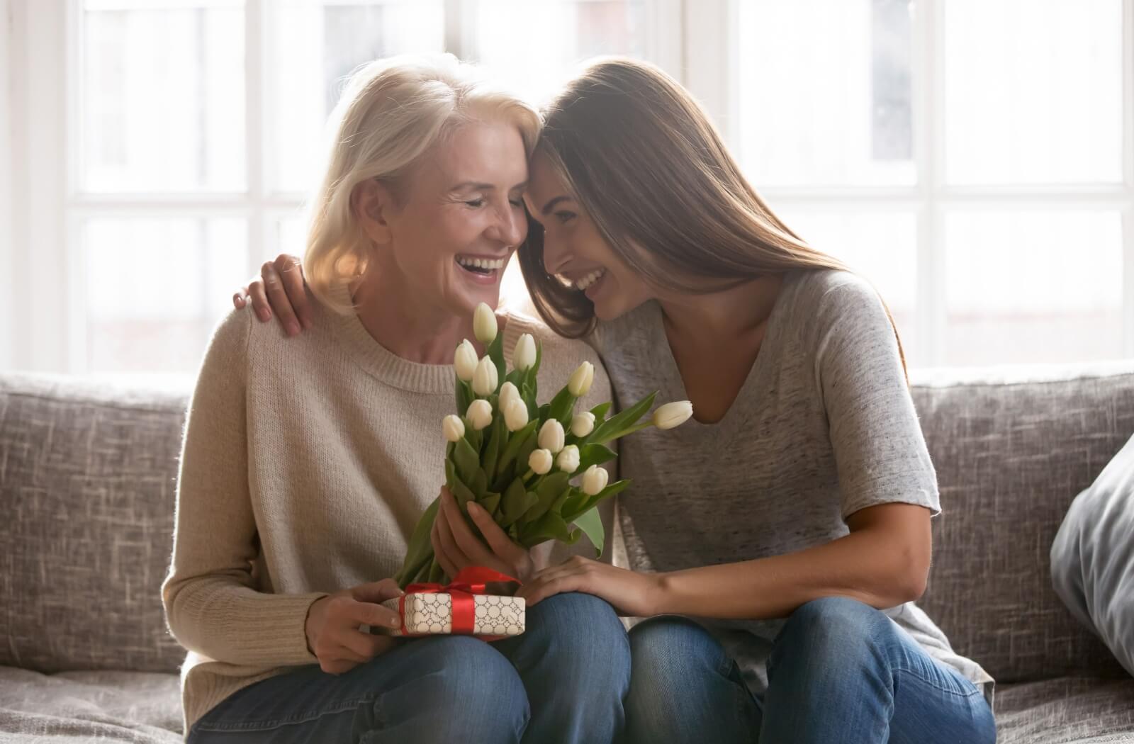 An adult child hugs their parent after giving them a bouquet of white roses and a present during a visit