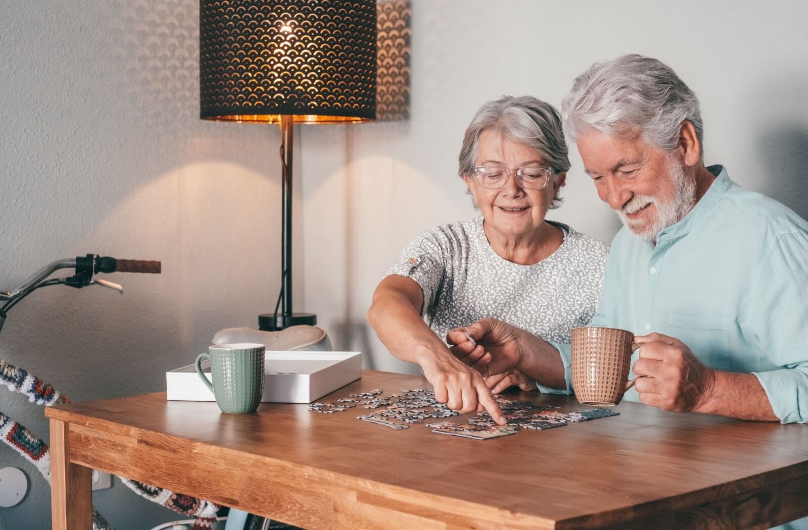 A pair of older adults sit in their living room and work together on a puzzle while drinking coffee and laughing