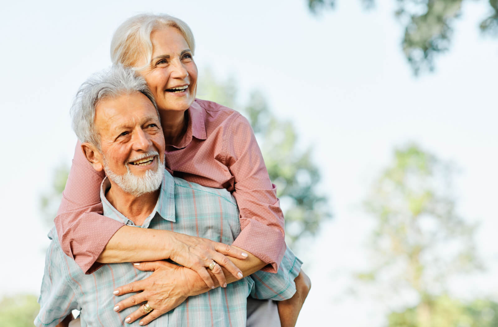 An older married couple smiling during a piggyback ride outside in the park.