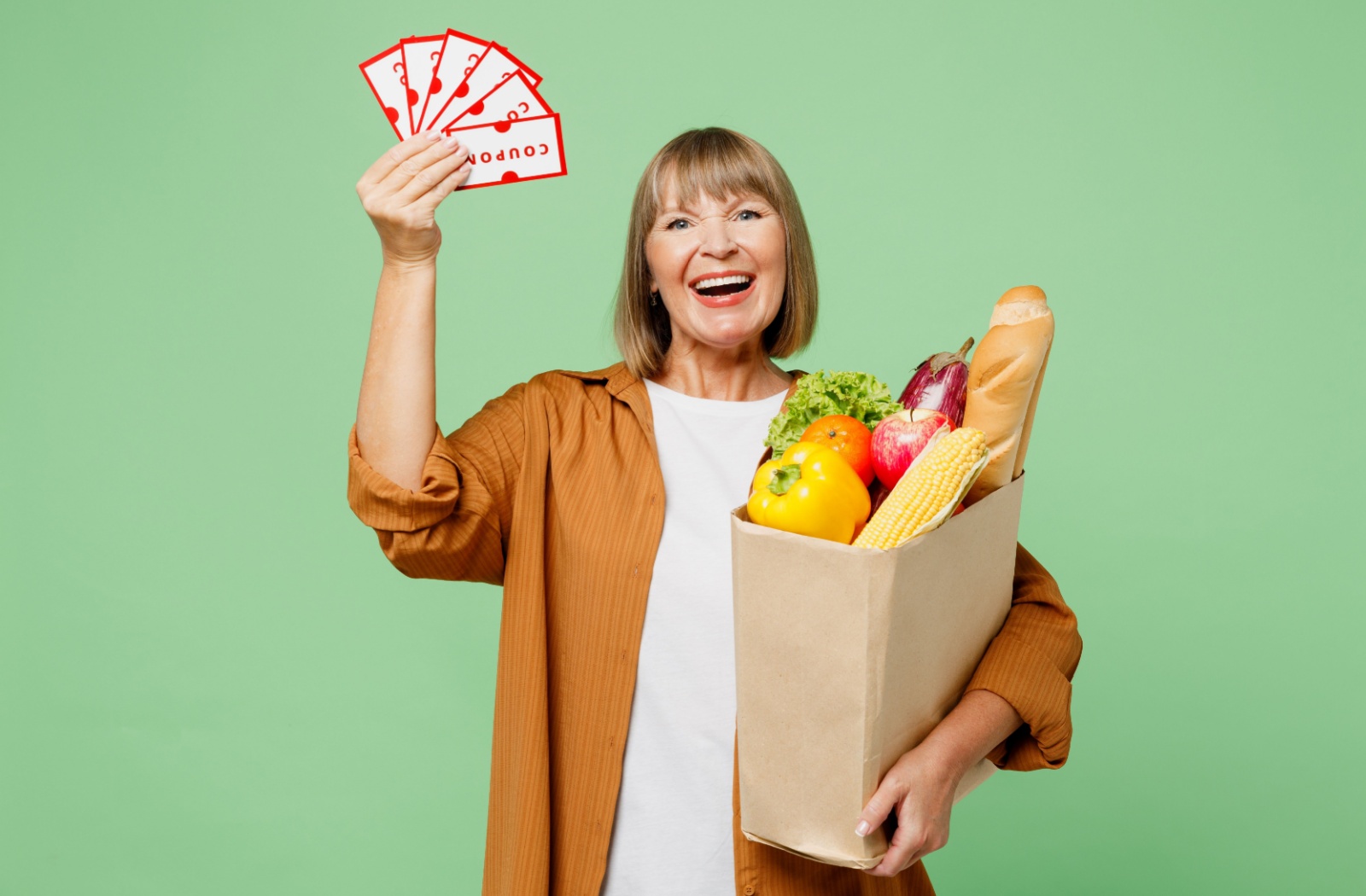 Against a green background, a happy senior holds coupons in one hand and a paper bag of groceries in the other