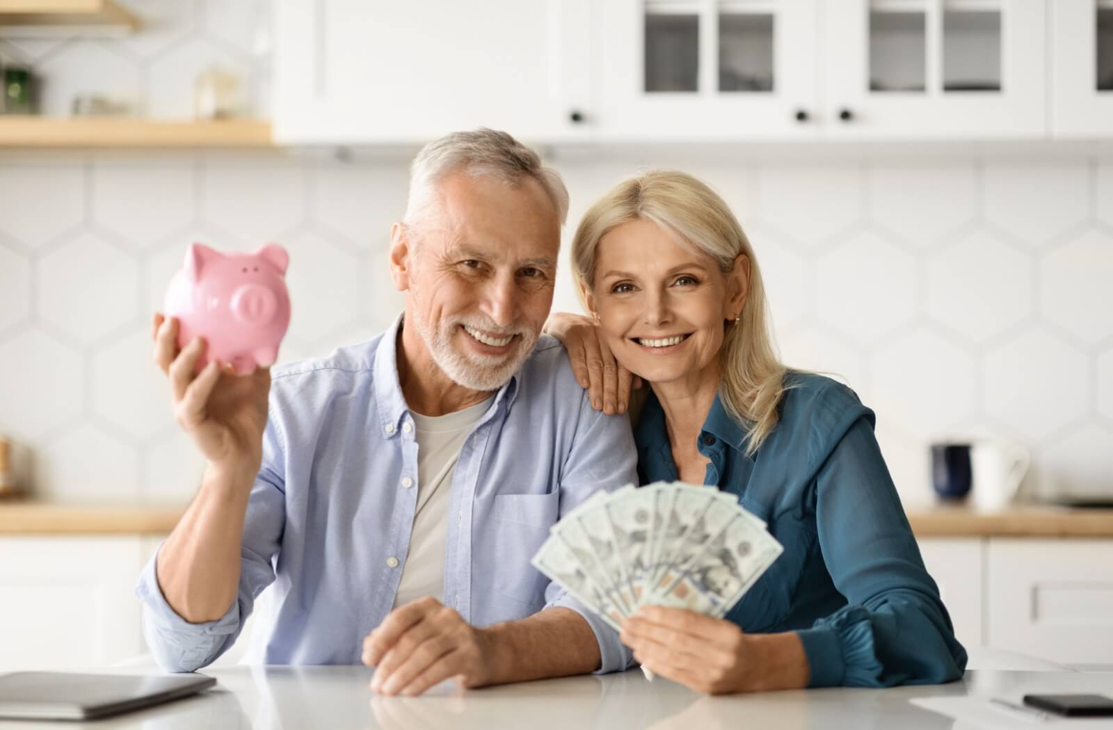 A smiling senior couple hold a pink piggy bank and a spread of US hundred-dollar bills to represent their savings