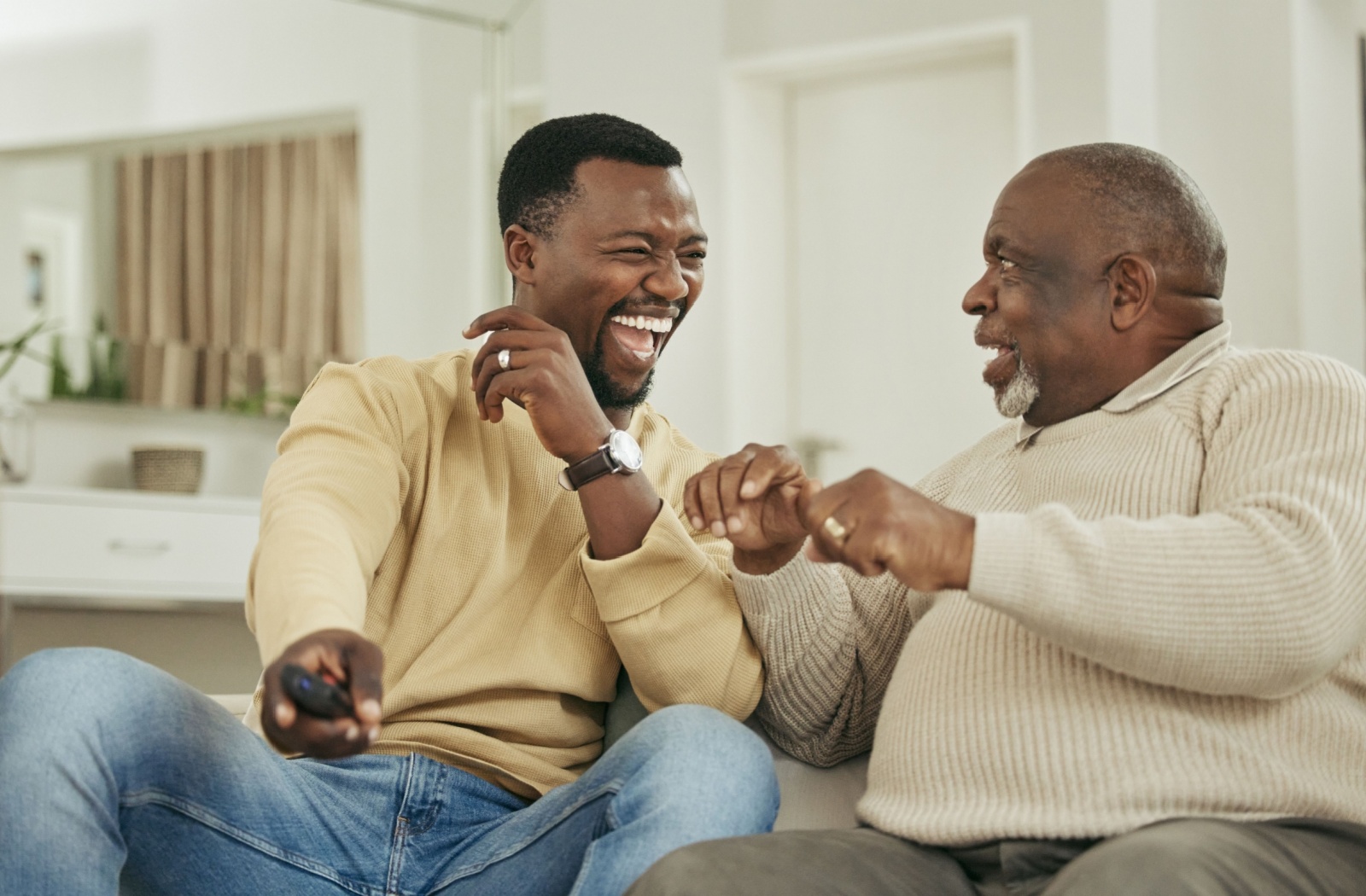 An adult child laughs while sitting on the couch with their older parent during a visit to their assisted living home