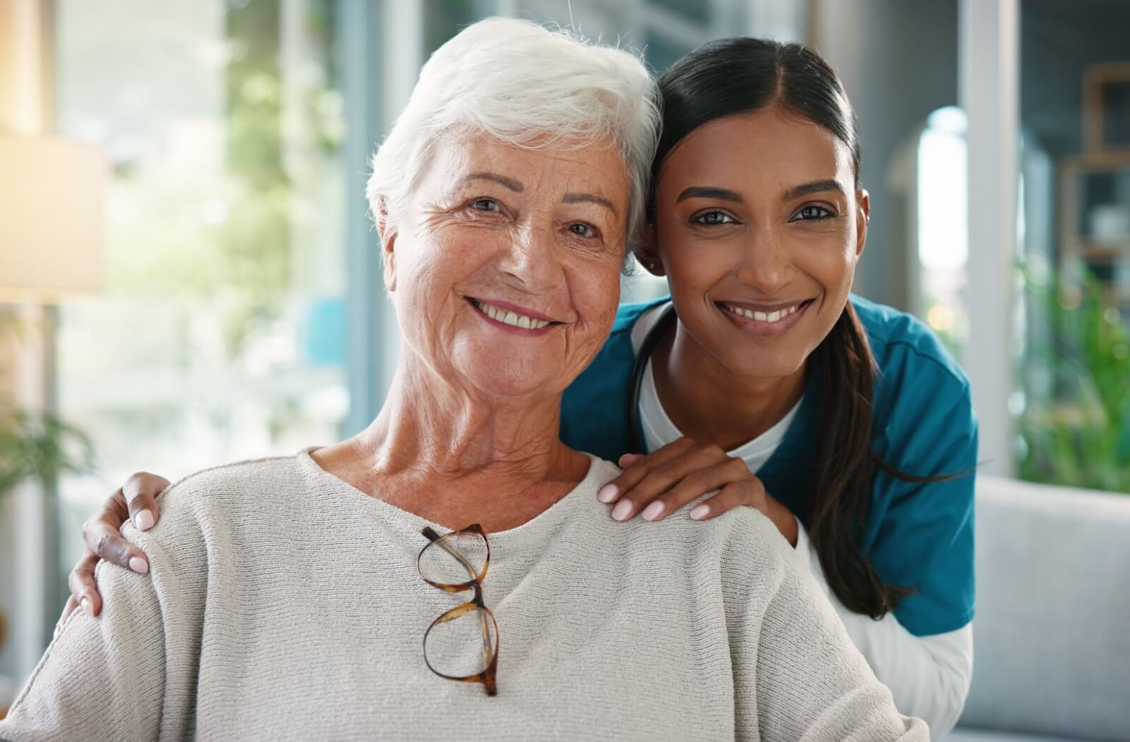 A smiling senior and a supportive caregiver in scrubs sit closely together in a bright living room.
