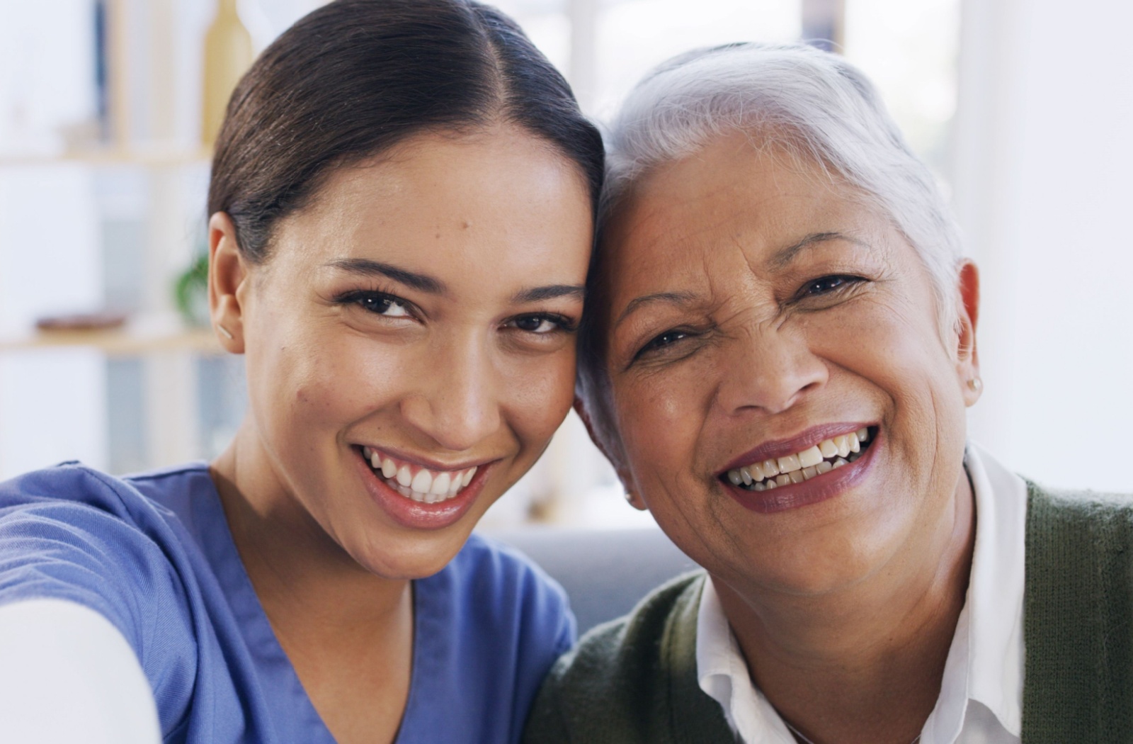 Cheerful caregiver in scrubs taking a close selfie with a senior woman smiling warmly in a cozy indoor setting