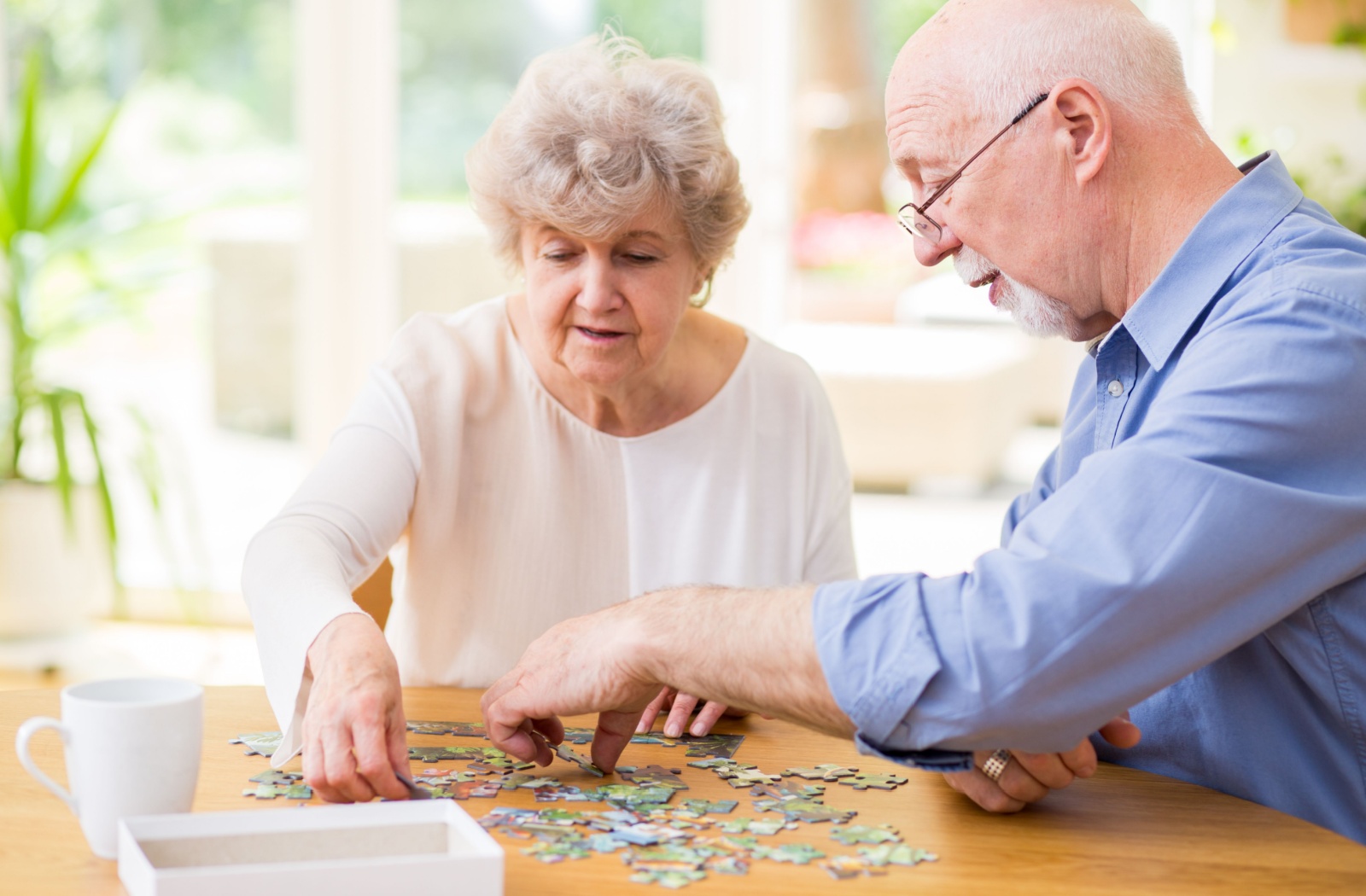 Two older adults smile in a sunlit lounge while assembling a puzzle over mid-afternoon tea