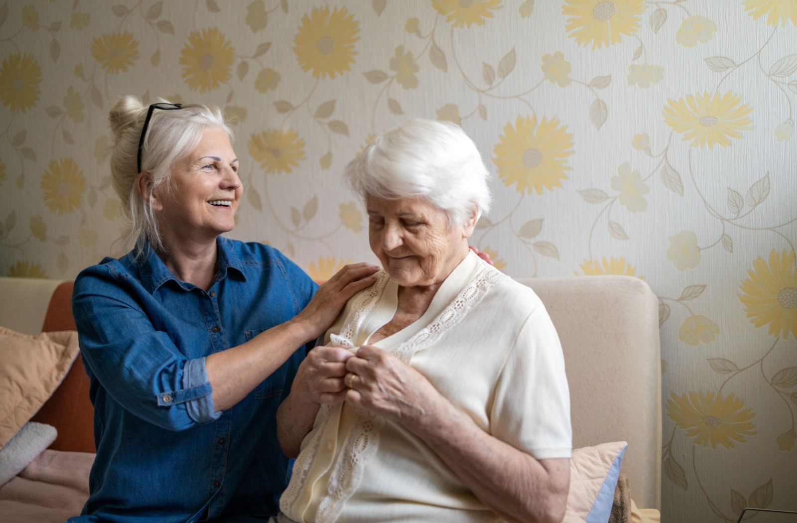 An adult child smiles as they assist their parent with dementia as they button their blouse.
